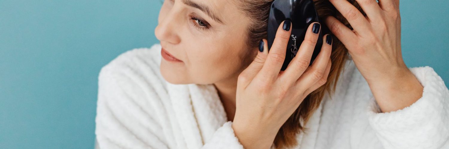 Vrouw in witte badjas borstelt haar lange haar voor de spiegel als onderdeel van haar haarverzorging