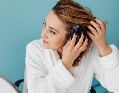 Vrouw in witte badjas borstelt haar lange haar voor de spiegel als onderdeel van haar haarverzorging